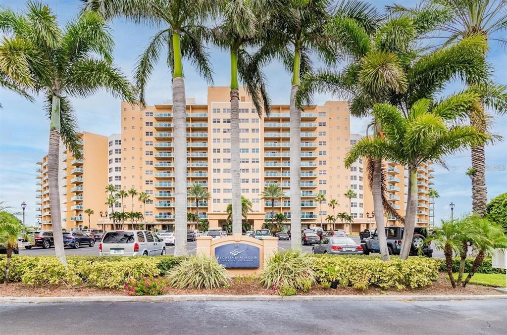 A multi-story tan apartment building stands behind a parking lot with palm trees and landscaping — 880 MANDALAY AVENUE UNIT S806, CLEARWATER BEACH, FL