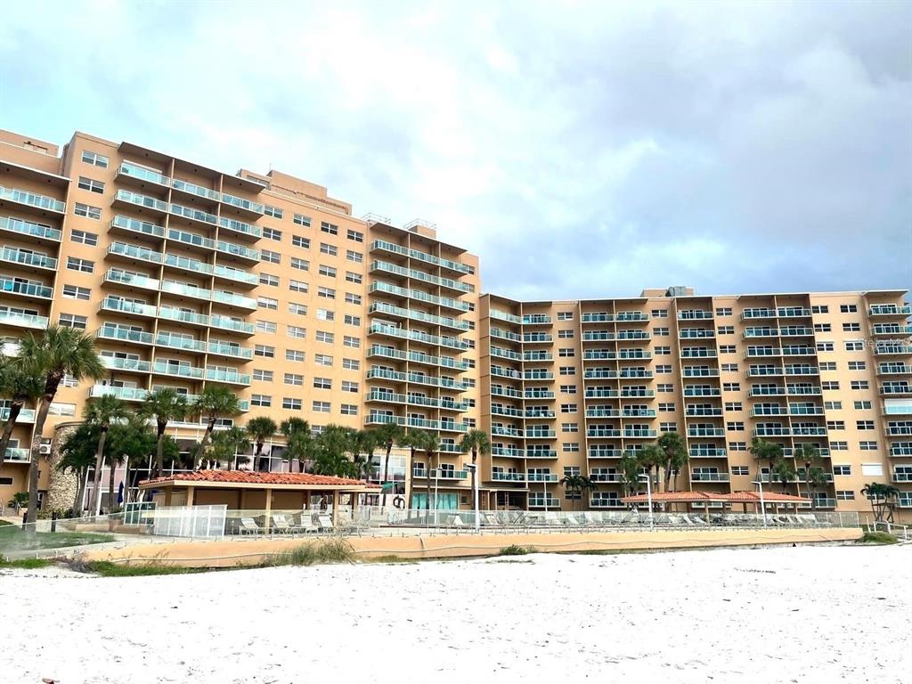 A large, multi-story tan apartment building stands behind a sandy beach and pool area — 880 MANDALAY AVENUE UNIT S806, CLEARWATER BEACH, FL