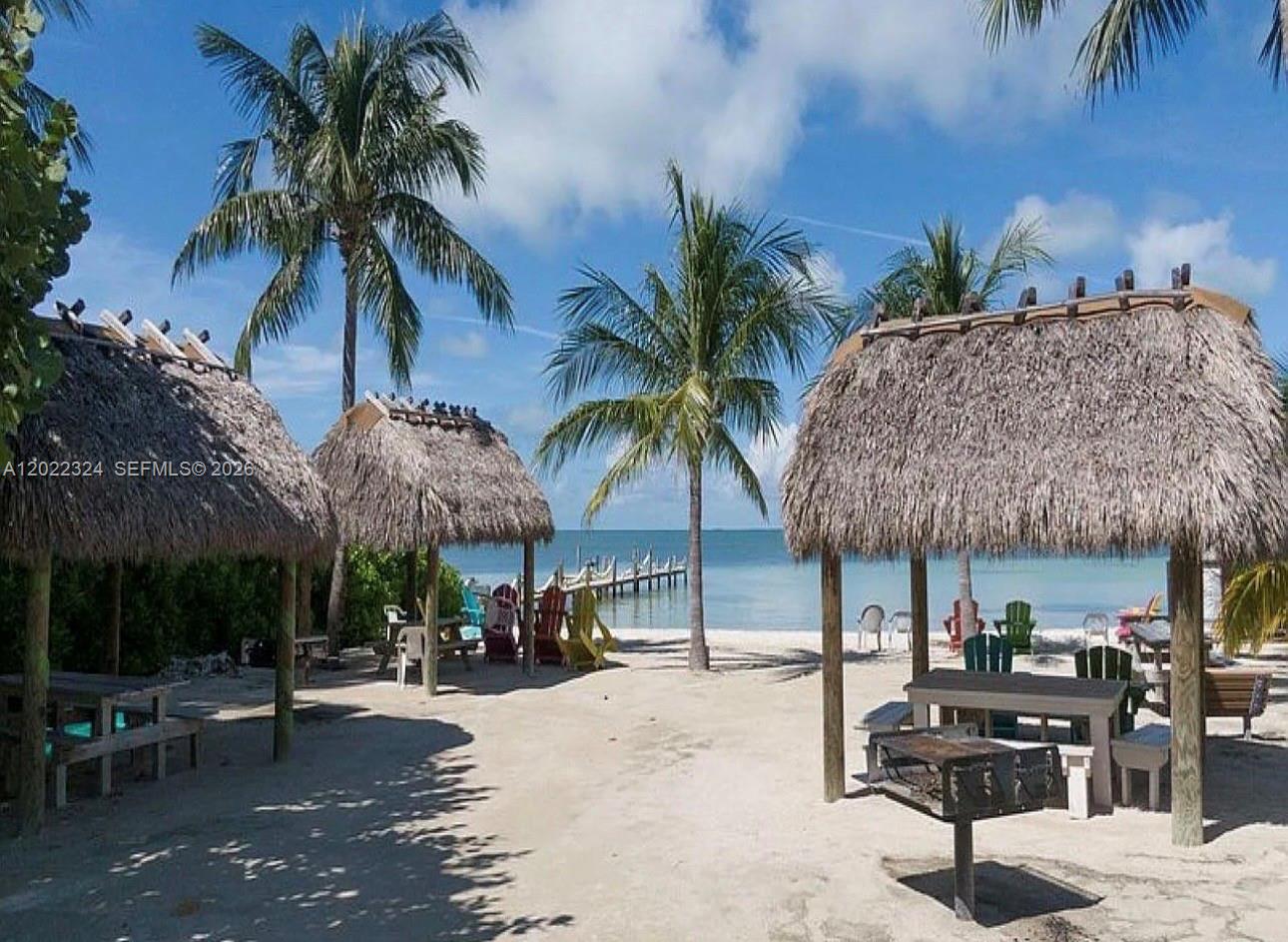 A sandy beach area features three thatched-roof tiki huts, palm trees, and a wooden pier — 102 Venetian Dr # 102, Islamorada FL 33036, ISLAMORADA, FL