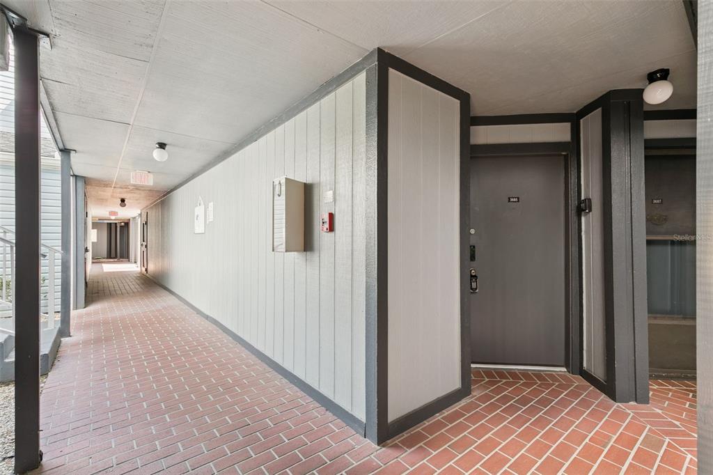 An outdoor apartment hallway features brick flooring, light-colored siding, and dark-trimmed entry doors — 3685 ATRIUM DRIVE UNIT L1, ORLANDO, FL