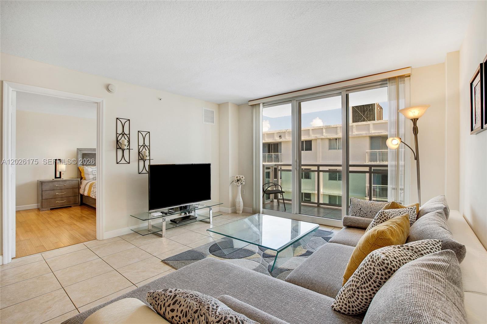 A living room features a gray sectional sofa, glass coffee table, and sliding glass balcony doors — 6917 Collins Ave # 1608, Miami Beach FL 33141, MIAMI BEACH, FL
