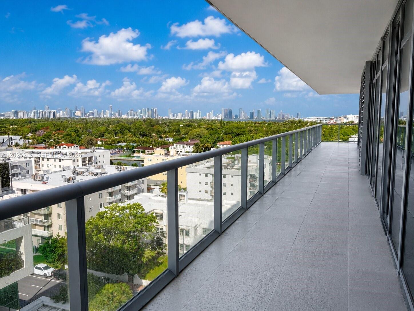 A spacious balcony with glass railings overlooks a lush neighborhood and a distant city skyline — 3737 Collins Ave # 1003, Miami Beach FL 33140, MIAMI BEACH, FL