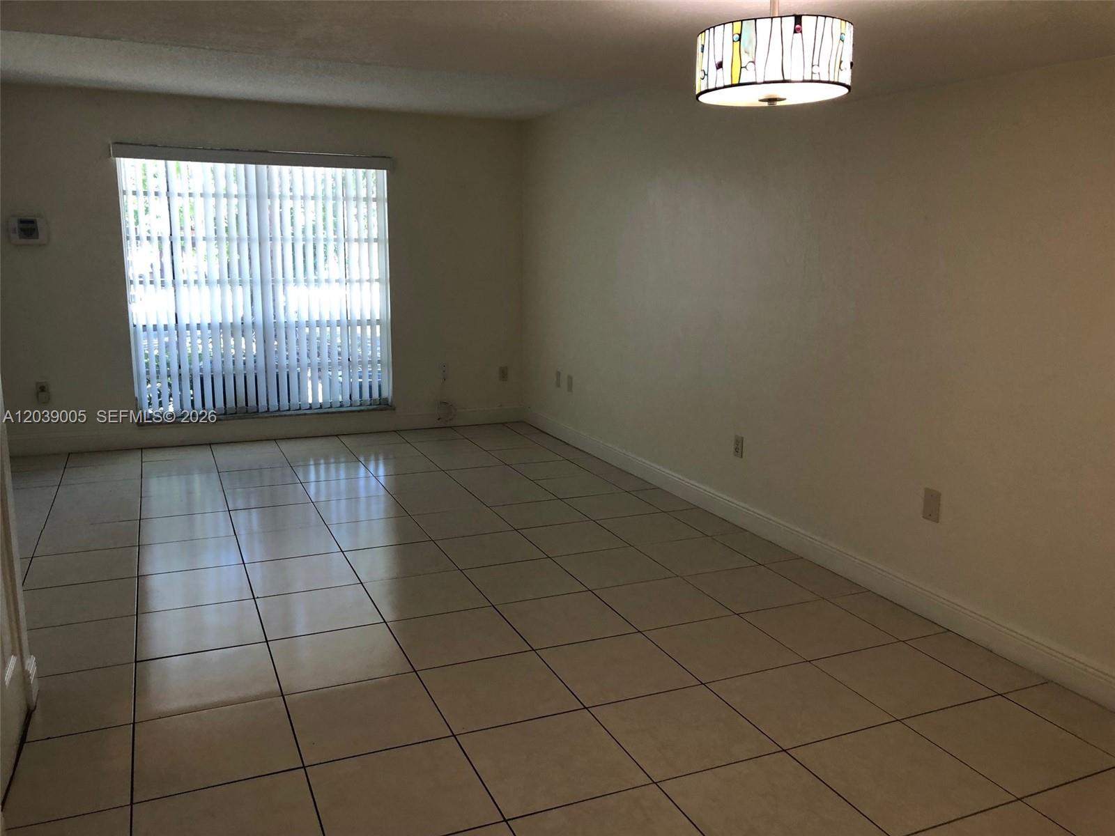 An empty room features beige tiled flooring, white walls, a large window, and a decorative light — 10808 N Kendall Dr # 4, Miami FL 33176, MIAMI, FL
