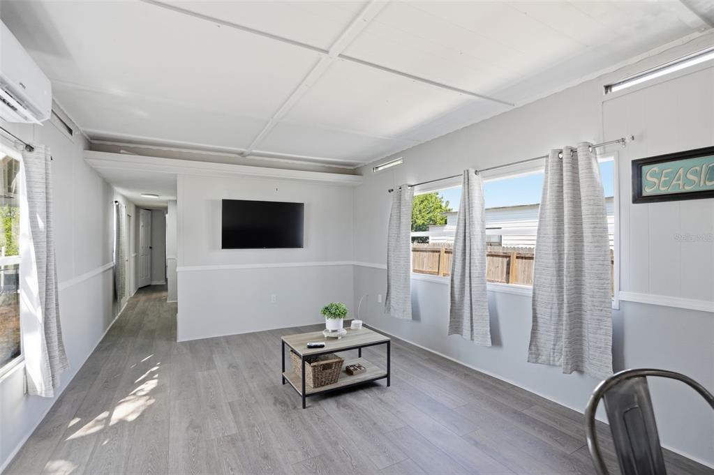 A living room features light gray wood-look flooring, white walls, a wall-mounted television, and windows — 6727 ALBATROSS LANE, HUDSON, FL