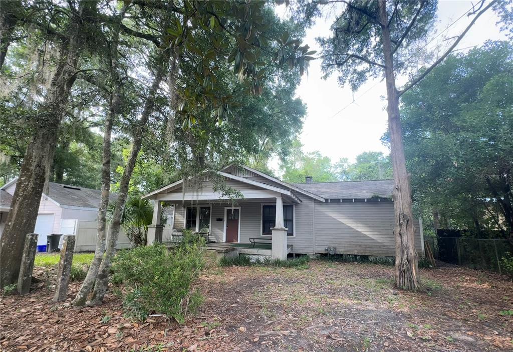 A single-story house with gray siding and a front porch sits among mature trees — 907 NW 11TH AVENUE, GAINESVILLE, FL