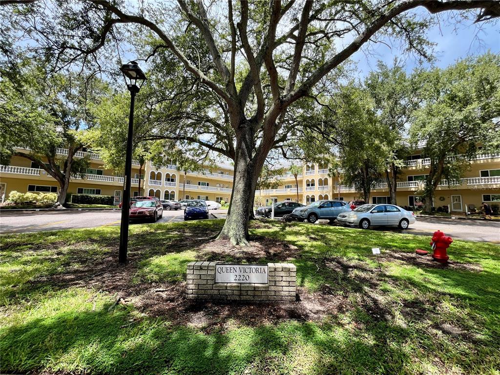 A multi-story apartment building sits behind a large tree and a brick sign — 2220 SPANISH DRIVE UNIT 54, CLEARWATER, FL