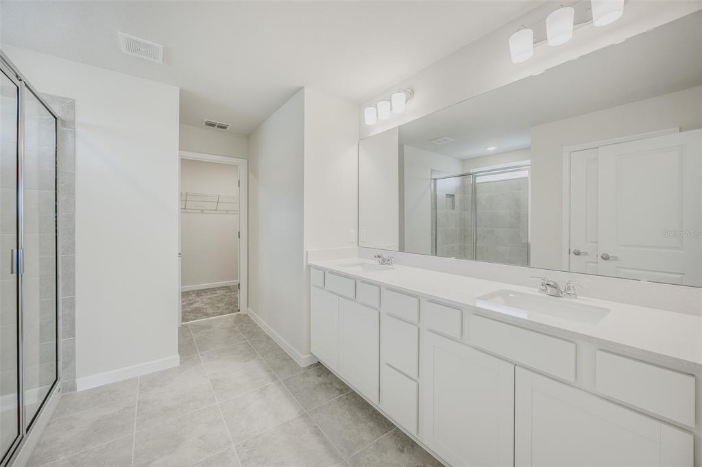A modern bathroom features a double vanity with white cabinets, gray tile flooring, and a shower — 6090 WINDY LEAF ALLEY, ST CLOUD, FL