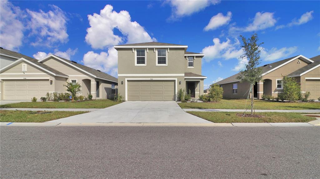 A two-story house with a tan stucco exterior and a two-car garage faces the street — 5546 LOGAN CAVE AVENUE, WIMAUMA, FL