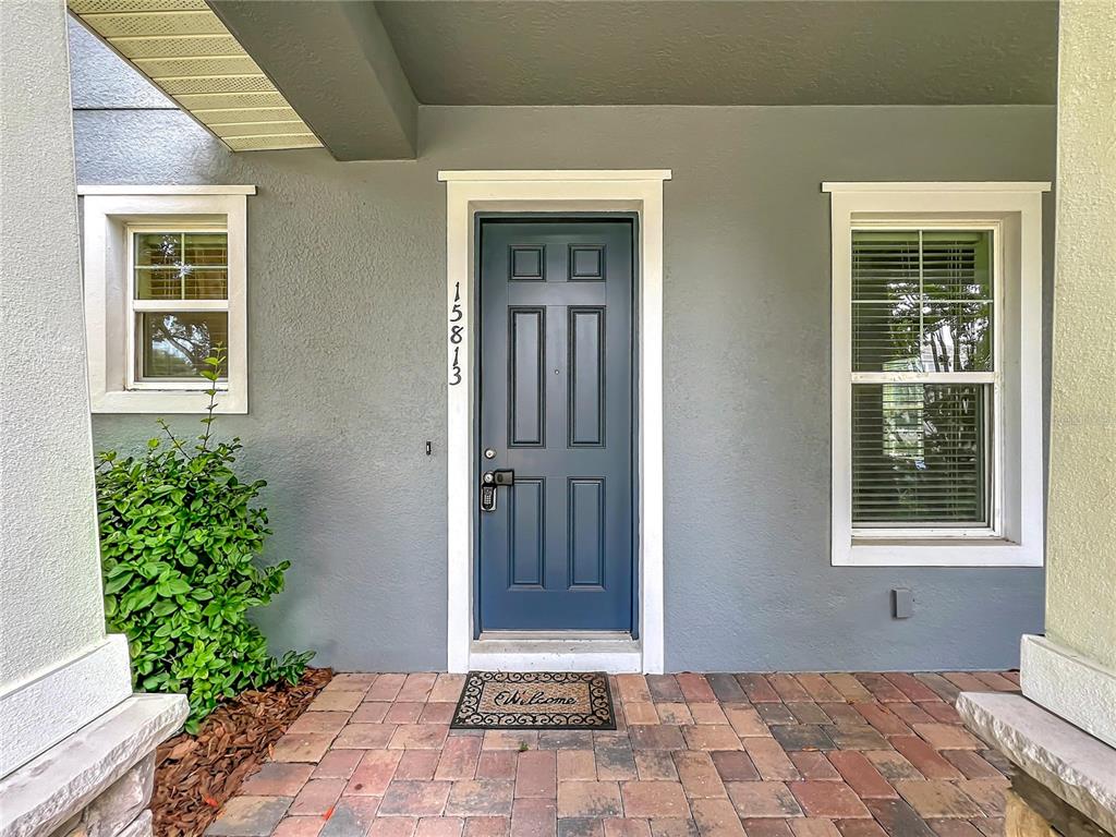 A blue front door with a welcome mat sits between two windows on a porch — 15813 SWEET LEMON WAY, WINTER GARDEN, FL