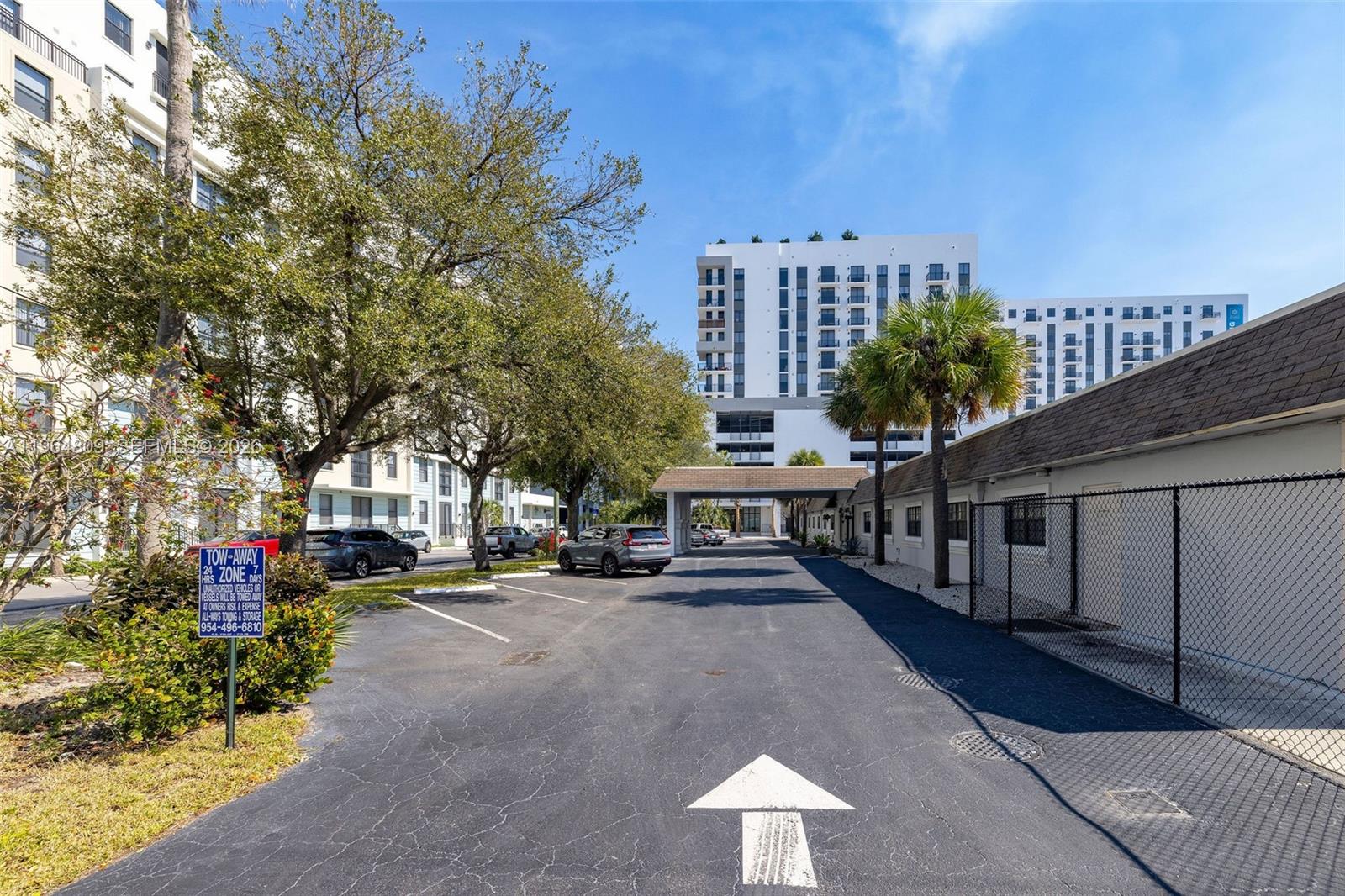An outdoor parking lot leads toward a modern multi-story apartment building under a blue sky — 151 SW 1st St, Dania Beach FL 33004, DANIA BEACH, FL