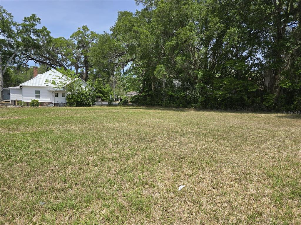A grassy, vacant lot sits in front of a white house surrounded by mature trees — 33 NW 3RD STREET, WILLISTON, FL