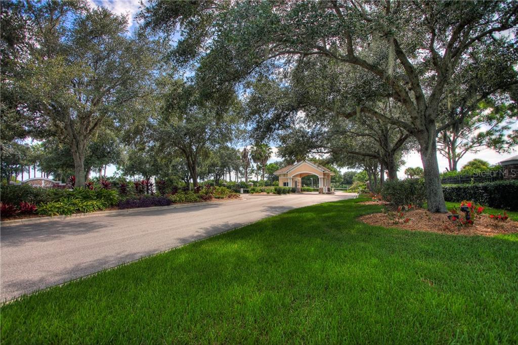 A paved road leads toward a gated community entrance under a canopy of large trees — 5330 HYLAND HILLS AVENUE UNIT 2321, SARASOTA, FL