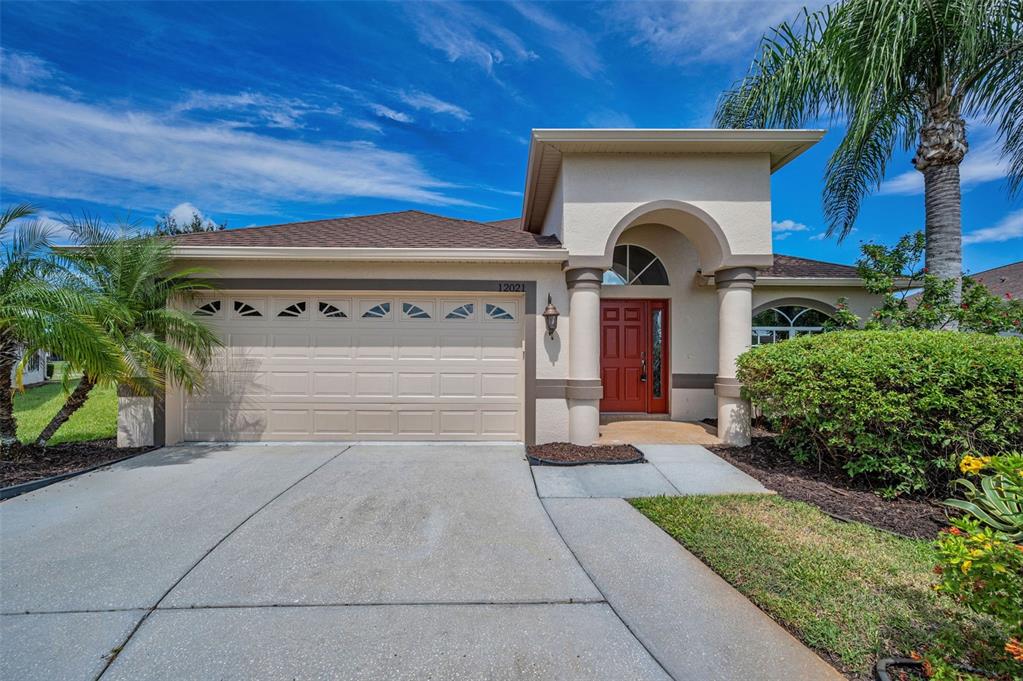 A single-story house with a beige stucco exterior, a two-car garage, and a red front door — 12021 FERN TERRACE COURT, TRINITY, FL