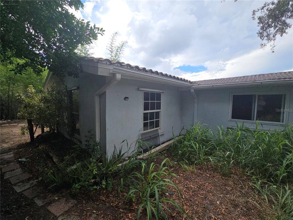 The exterior of a light-colored house features a tiled roof, gutters, and overgrown landscaping — 7809 WAXWOOD DRIVE, PORT RICHEY, FL