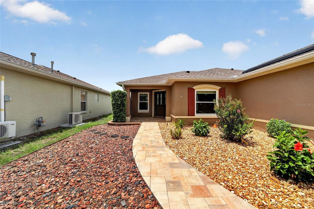 A paved walkway leads to the front entrance of a tan house with rock landscaping — 9702 SW 92ND PLACE ROAD, OCALA, FL