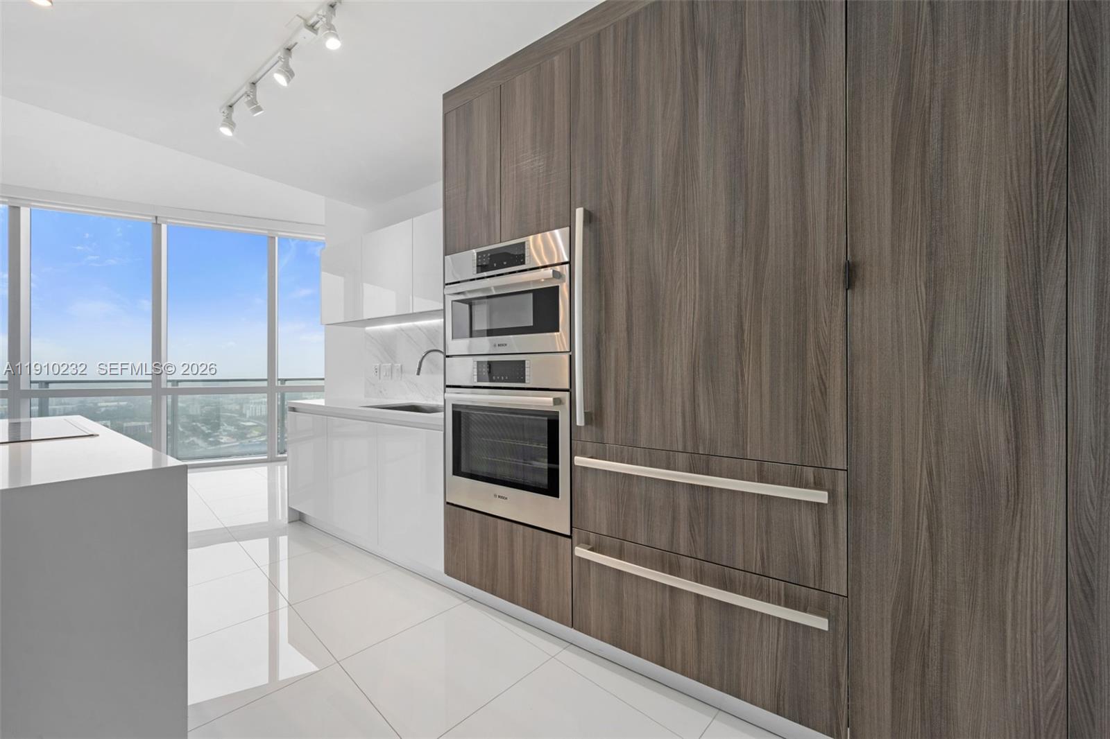 A modern kitchen features white glossy cabinets, wood-paneled appliances, and floor-to-ceiling windows with views — 851 NE 1st Ave # 4204, Miami FL 33132, MIAMI, FL