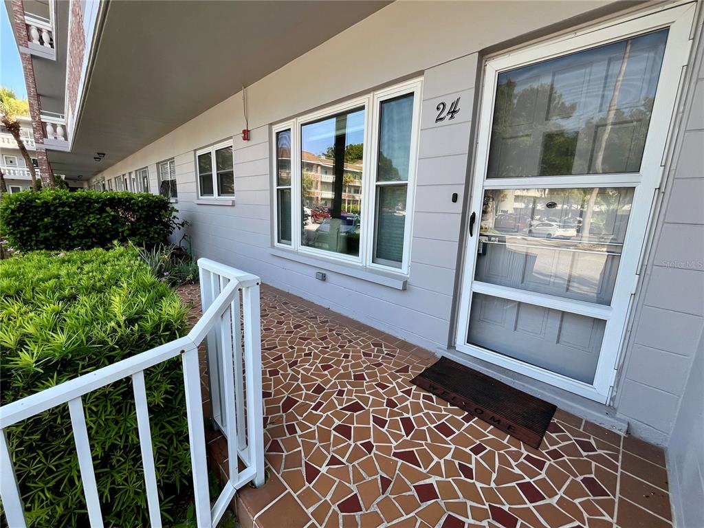 A ground-level apartment entrance features a tiled patio, a white door, and a white railing — 2460 PERSIAN DRIVE UNIT 24, CLEARWATER, FL