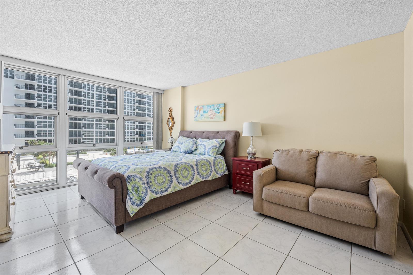 A bedroom features a gray tufted bed, a beige loveseat, and large windows overlooking buildings — 525 N Ocean Boulevard # 616, Pompano Beach FL 33062, POMPANO BEACH, FL