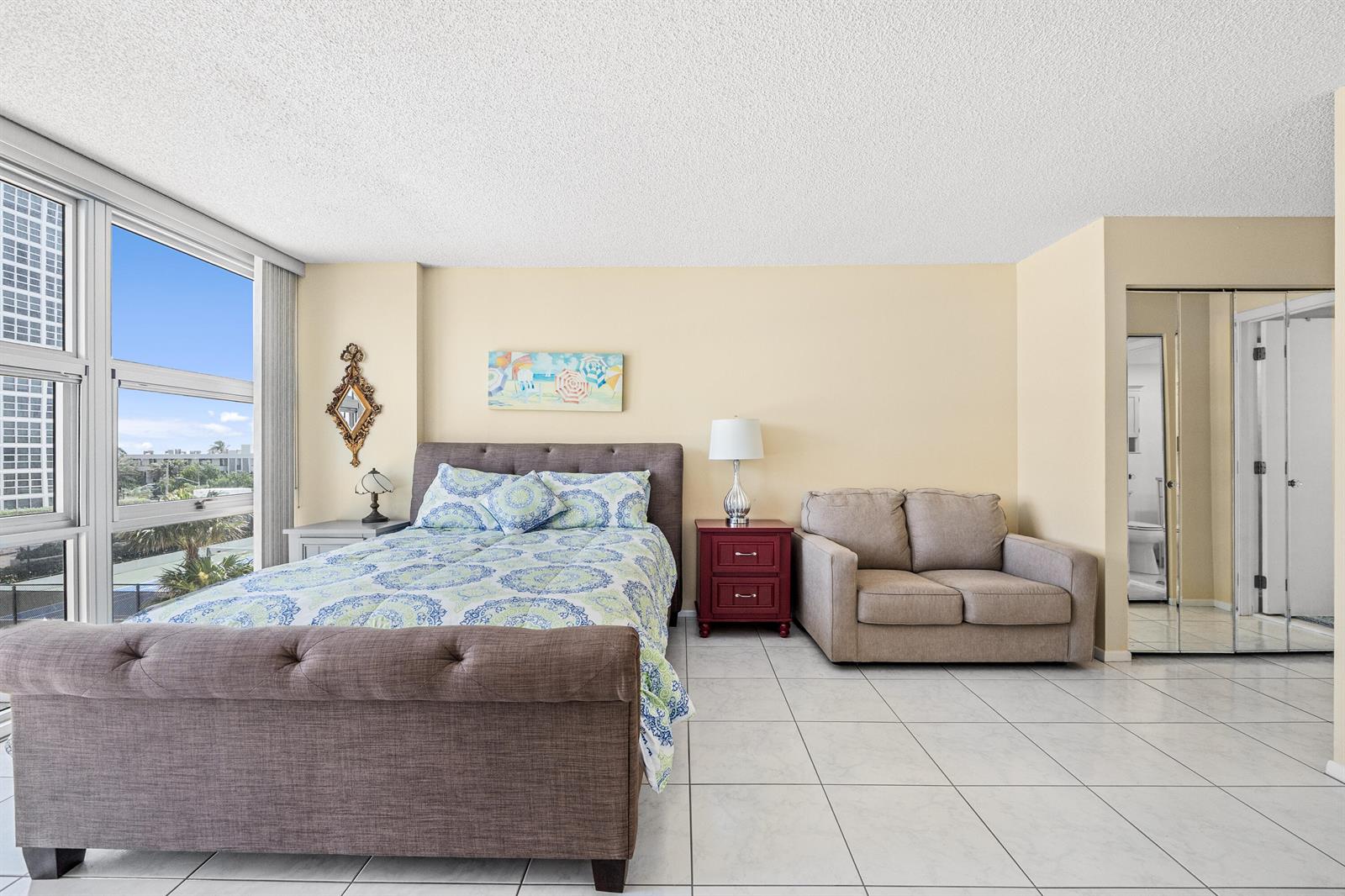 A bedroom features a tufted bed, a small sofa, a nightstand, and a large window — 525 N Ocean Boulevard # 616, Pompano Beach FL 33062, POMPANO BEACH, FL