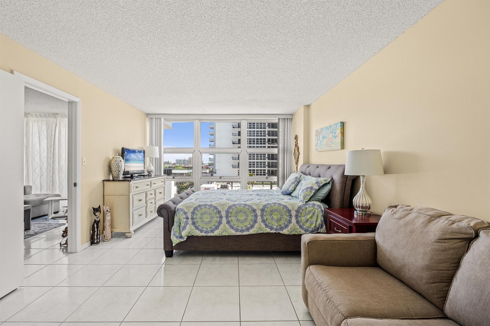 A bedroom features a bed, dresser, sofa, and large windows overlooking a city skyline — 525 N Ocean Boulevard # 616, Pompano Beach FL 33062, POMPANO BEACH, FL