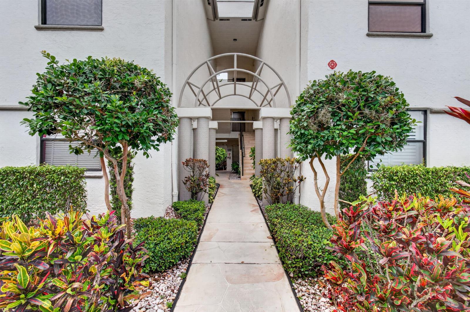 A paved walkway leads to the arched entrance of a multi-story residential building with landscaping — 5133 Brisata Circle # B, Boynton Beach FL 33437, BOYNTON BEACH, FL