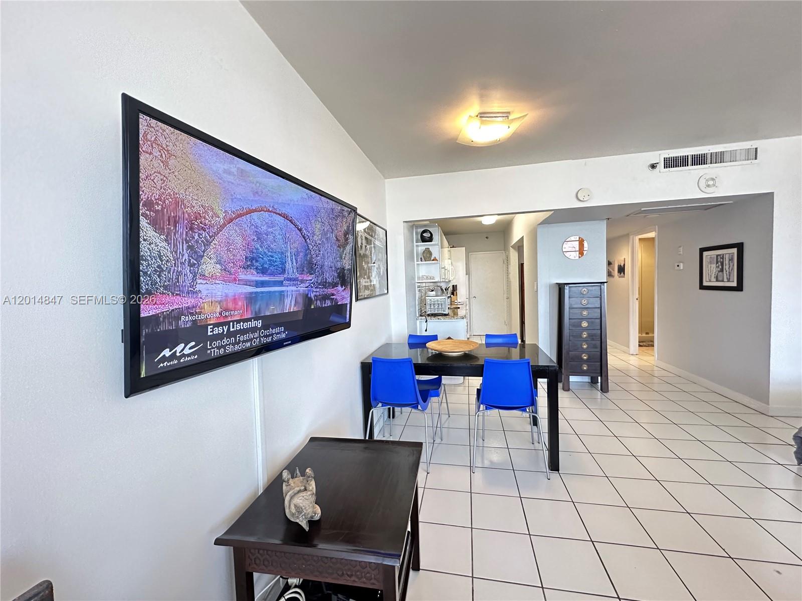 A dining area with a black table and blue chairs sits near a wall-mounted television — 100 Lincoln Rd # 1012, Miami Beach FL 33139, MIAMI BEACH, FL