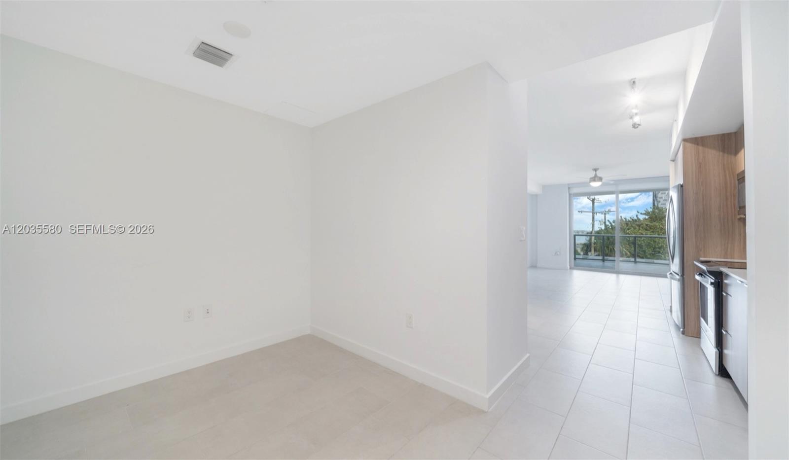 A bright, empty room with white walls and tile flooring leads to an open kitchen — MIAMI, FL
