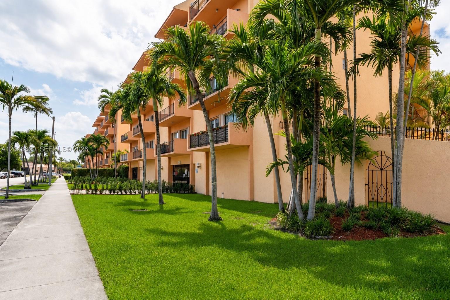 A multi-story peach-colored apartment building with balconies stands beside a sidewalk and palm trees — 1900 W 54th St # 220, Hialeah FL 33012, HIALEAH, FL