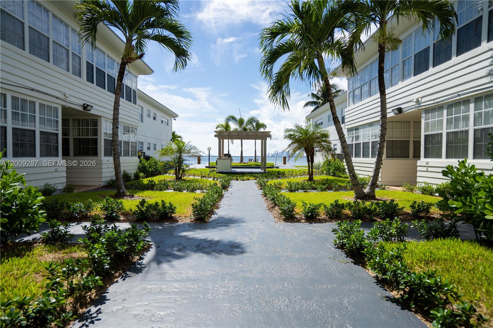 A paved walkway leads between two residential buildings toward a waterfront gazebo and palm trees — 1940 Bay Dr # 11, Miami Beach FL 33141, MIAMI BEACH, FL