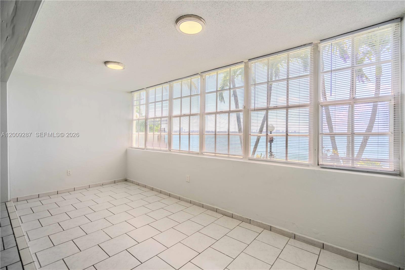 An empty room features white tile flooring, white walls, and large windows overlooking the water — 1940 Bay Dr # 11, Miami Beach FL 33141, MIAMI BEACH, FL