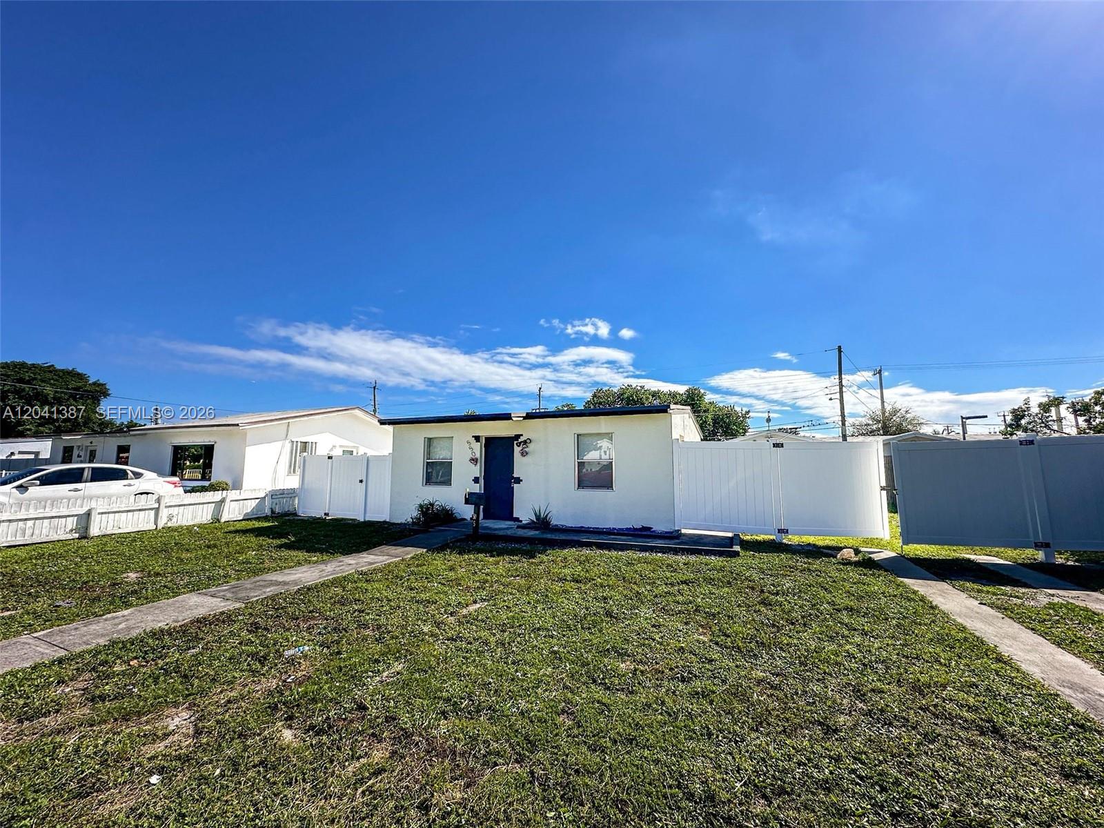 A white single-story house with a blue front door is enclosed by a white fence — 550 NW 15th Ct, Pompano Beach FL 33060, POMPANO BEACH, FL