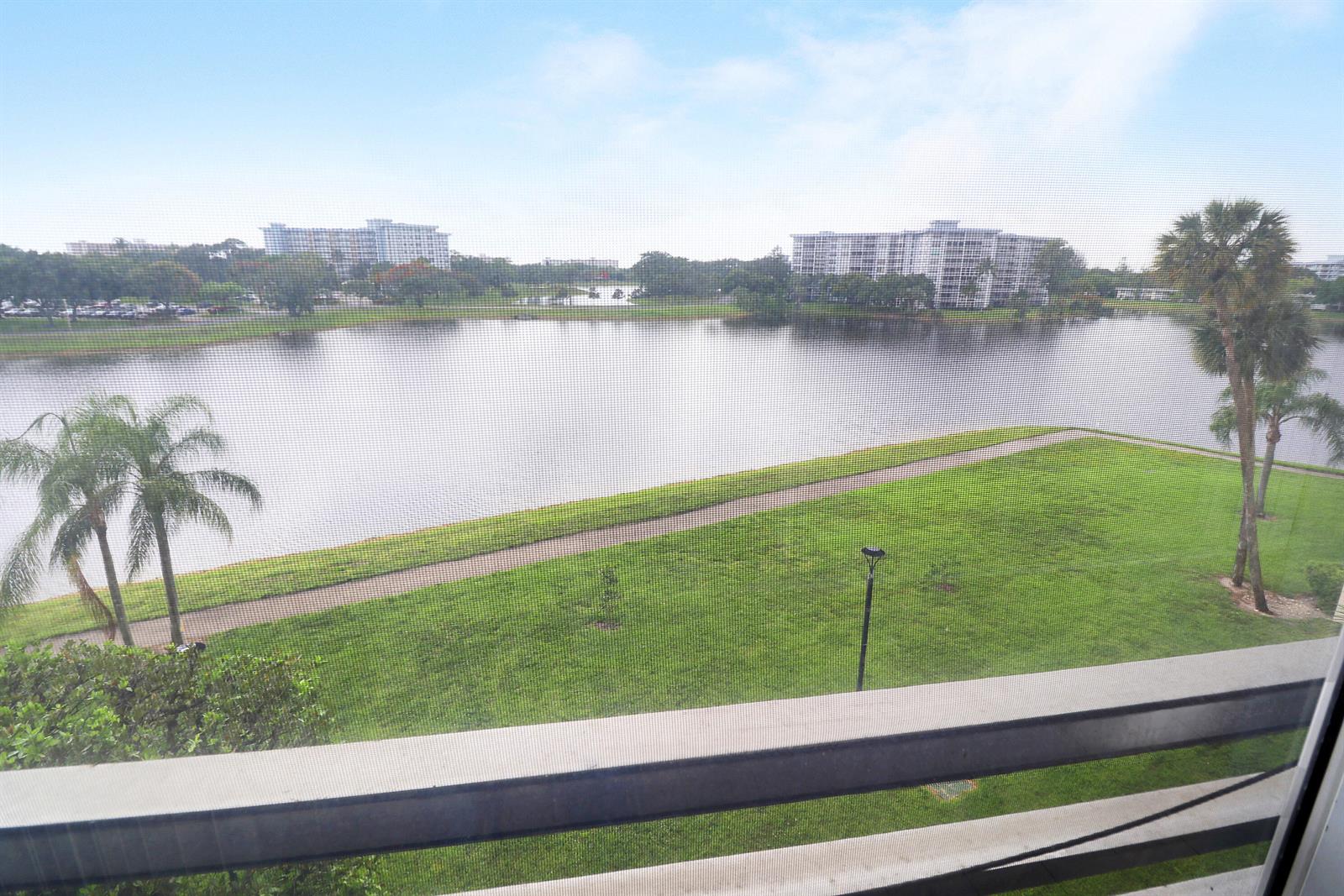 A view from a balcony overlooking a lake, green lawn, and distant apartment buildings — 3095 N Course Drive # 409, Pompano Beach FL 33069, POMPANO BEACH, FL
