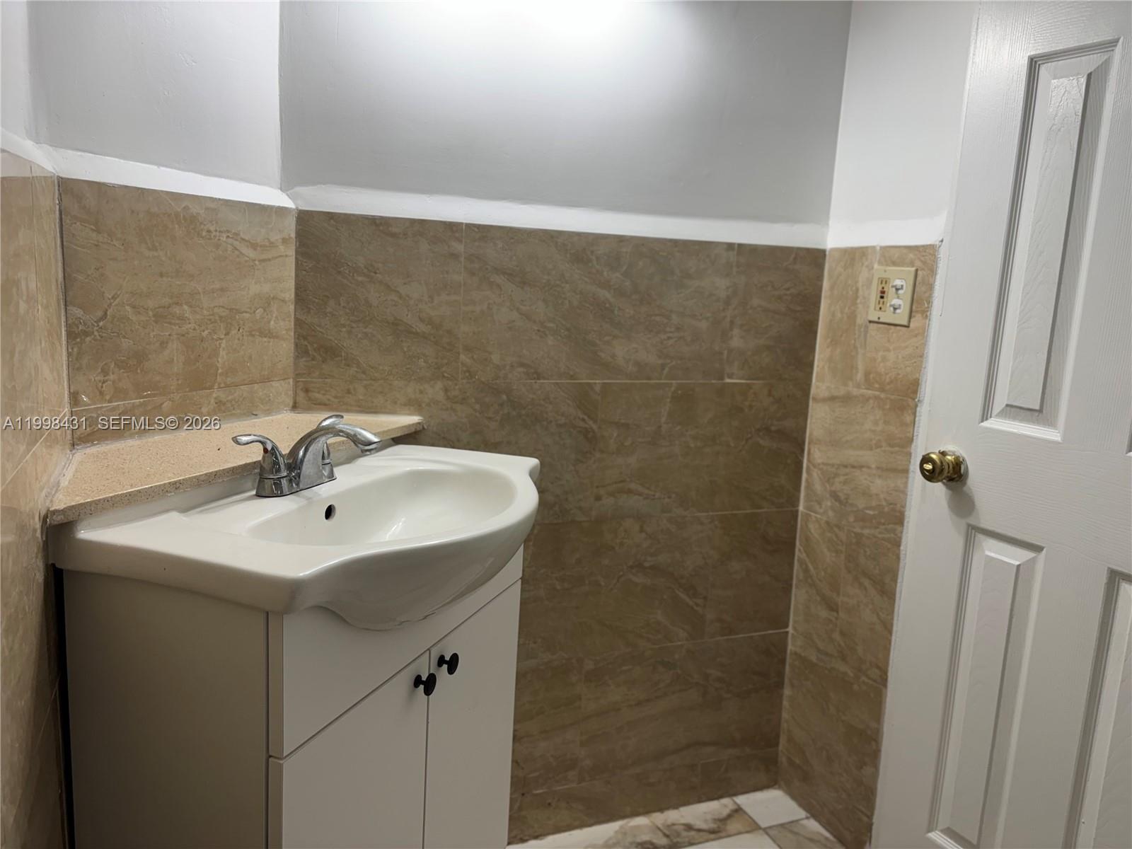 A bathroom features a white vanity with a sink, beige tiled walls, and a door — 1501 NE 191st St # C211, Miami FL 33179, MIAMI, FL