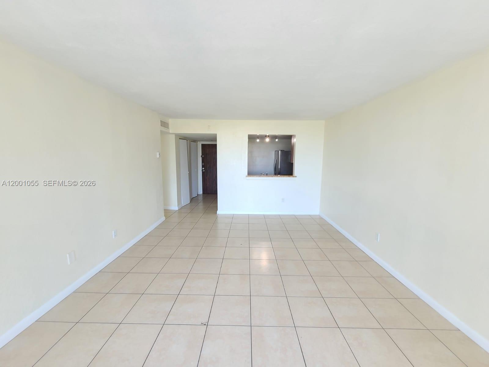 An empty living room features beige tile flooring, off-white walls, and a kitchen pass-through window — 496 NW 165th St Rd # 409, Miami FL 33169, MIAMI, FL