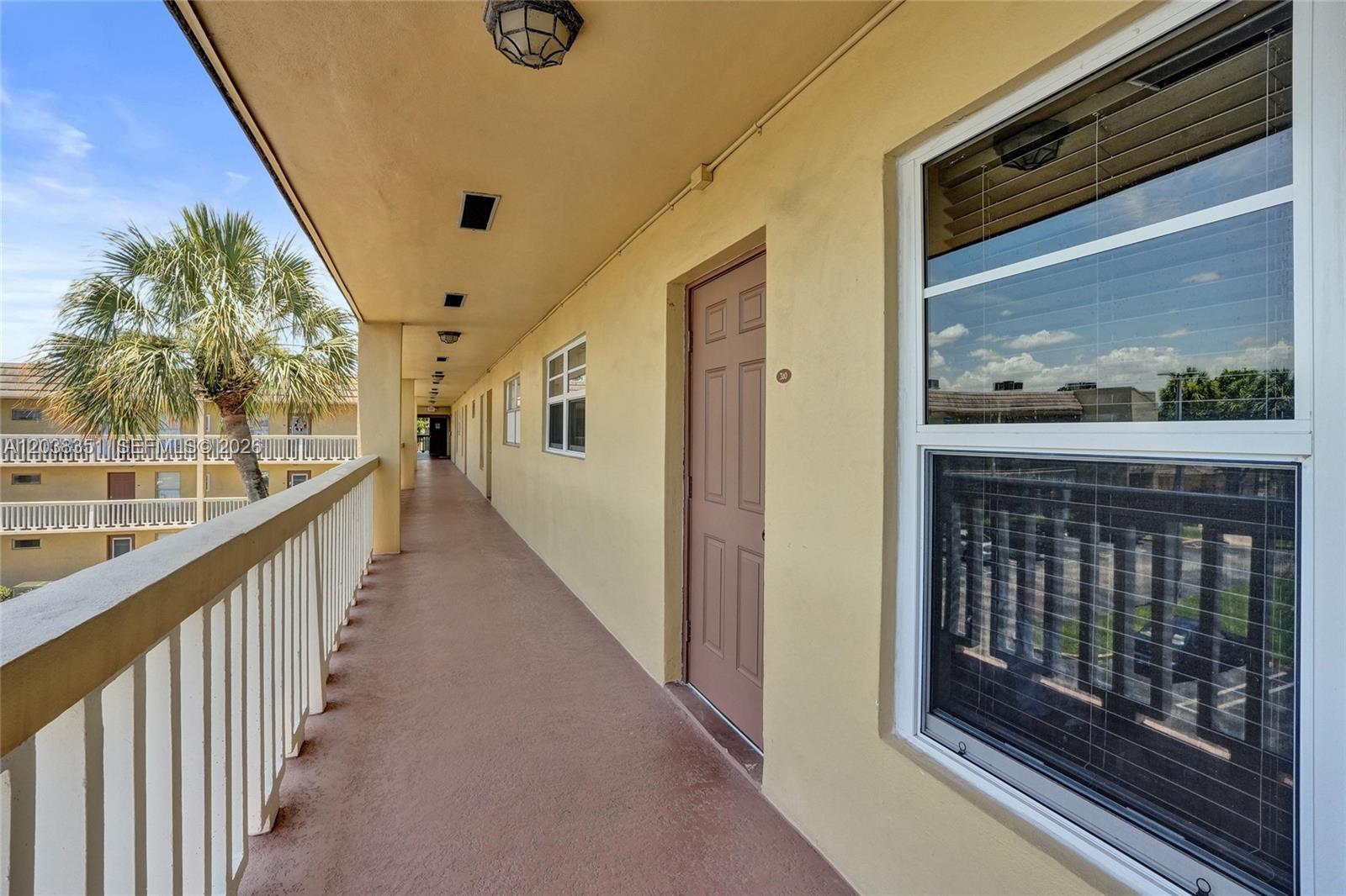 An exterior walkway with a brown door, white railing, and a view of palm trees — 5961 NW 61st Ave # 310, Tamarac FL 33319, TAMARAC, FL