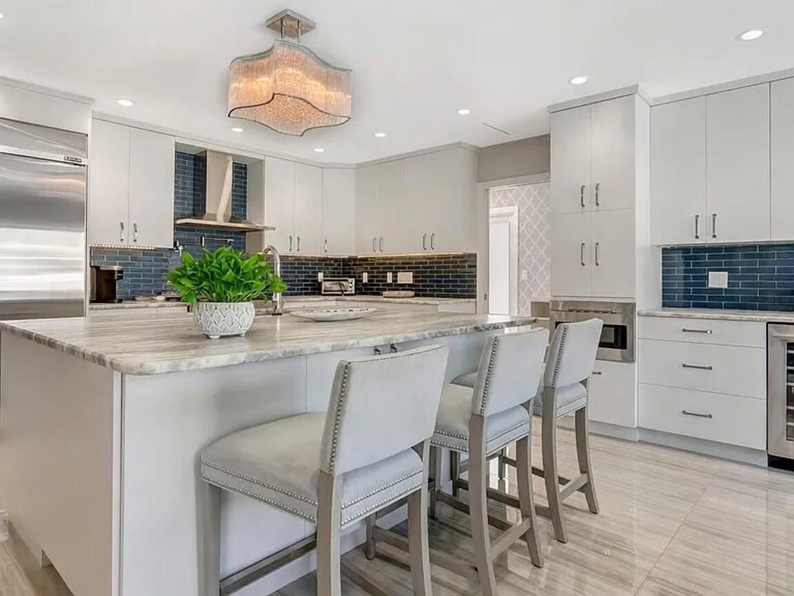 A modern kitchen features white cabinetry, a marble-topped island, bar stools, and blue tile backsplashes — 831 Dover Street, Boca Raton FL 33487, BOCA RATON, FL