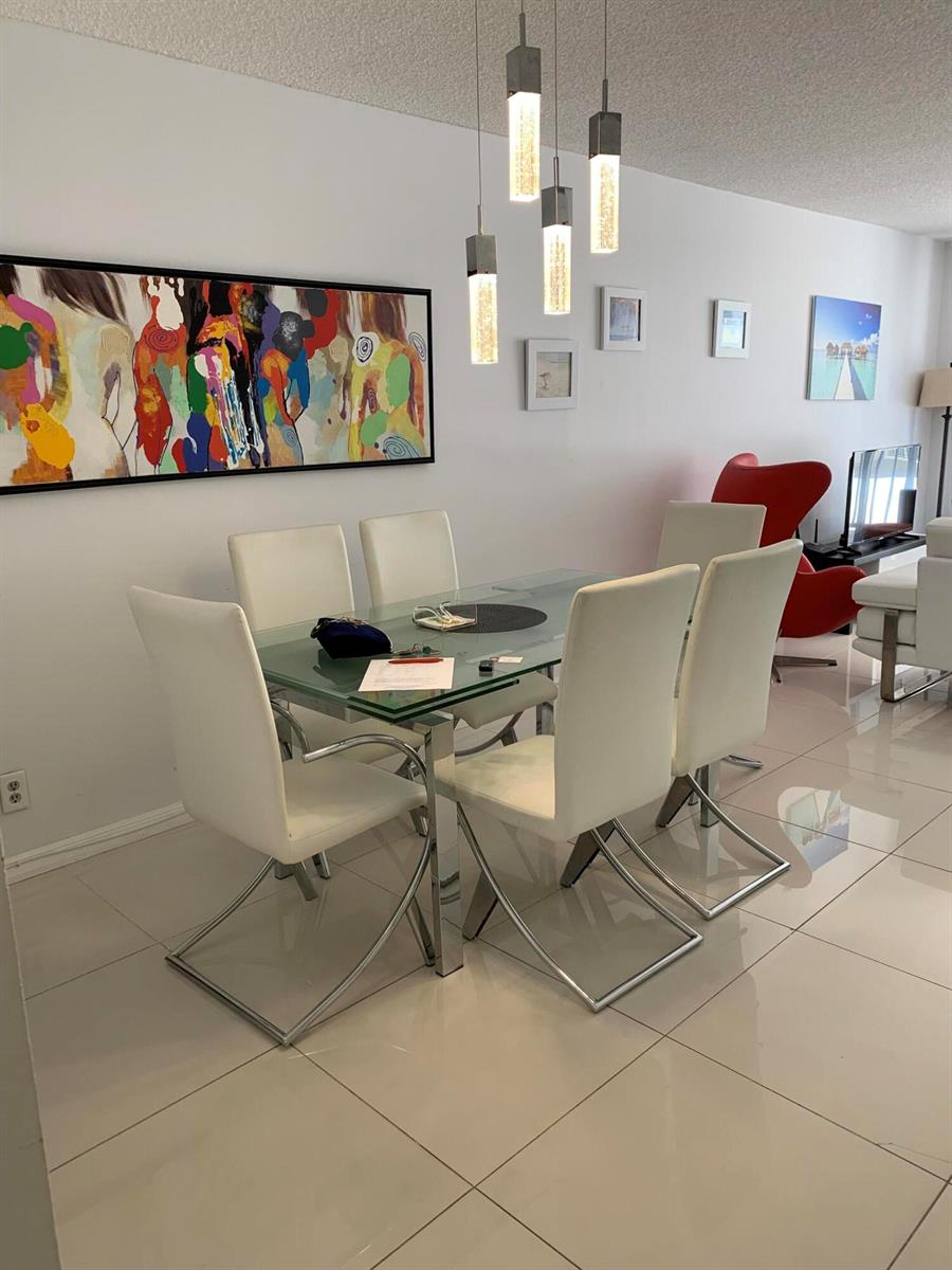 A dining room features a glass table, white chairs, modern pendant lights, and abstract wall art — 101 Briny Avenue # 806, Pompano Beach FL 33062, POMPANO BEACH, FL