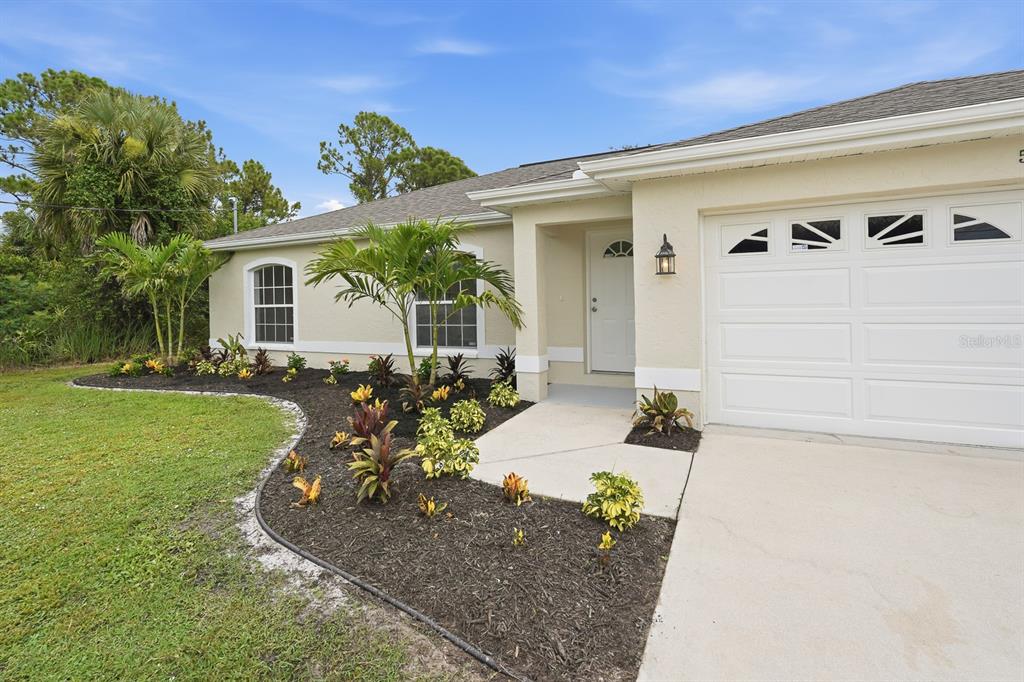 A single-story house exterior with a white garage door, front entrance, and landscaped garden bed — 5108 WEATHERTON STREET, NORTH PORT, FL
