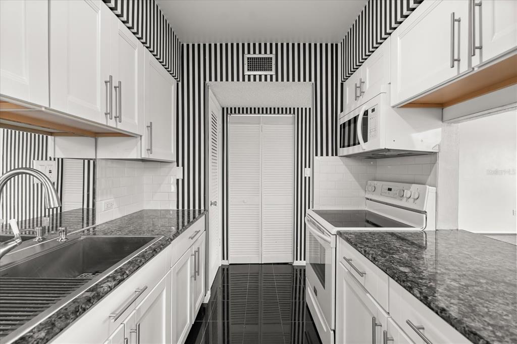 A galley kitchen features white cabinets, granite countertops, black-and-white striped wallpaper, and black tile flooring — 1700 6TH STREET NW UNIT B24, WINTER HAVEN, FL