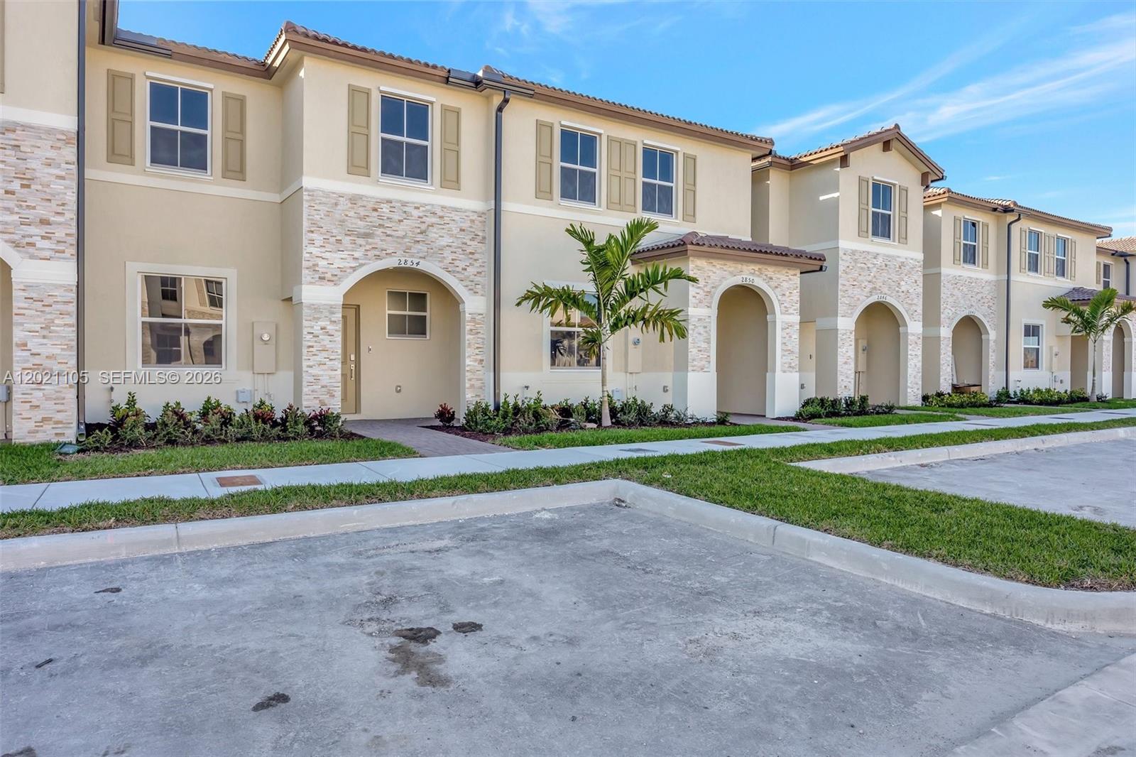 A row of two-story townhomes features beige stucco, stone accents, arched entryways, and paved parking — HOMESTEAD, FL
