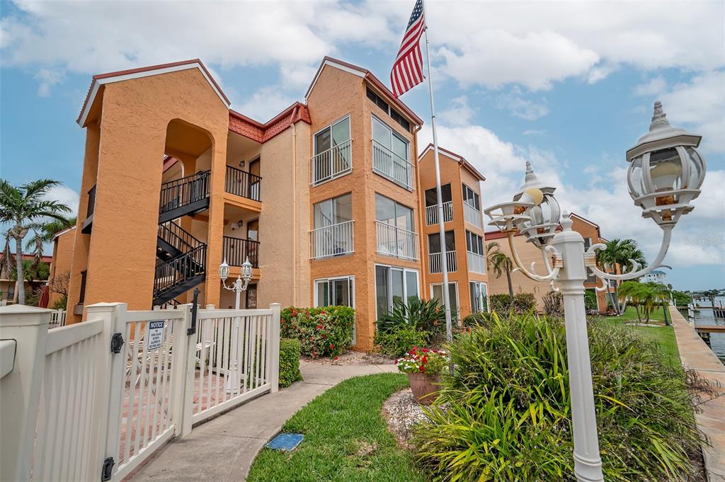 An exterior view of a multi-story orange apartment building with a white fence and flag — 8921 BLIND PASS ROAD UNIT 143, ST PETE BEACH, FL