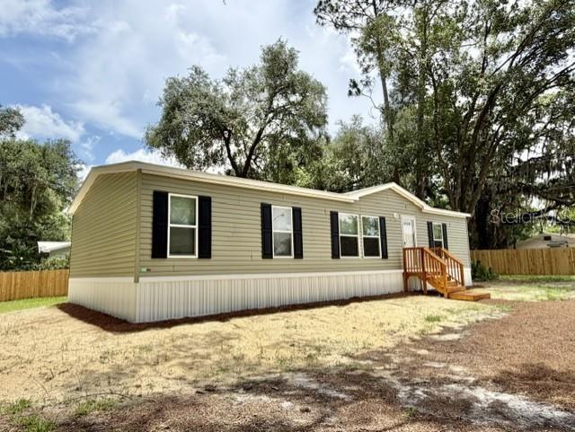 A light green manufactured home with black shutters stands on a dirt lot with trees — 713 NE 3RD STREET, CHIEFLAND, FL