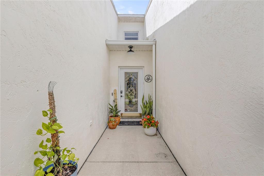 A narrow outdoor entryway features white stucco walls, a glass-paneled front door, and potted plants — 2236 DEERWOOD DRIVE, NEW SMYRNA BEACH, FL