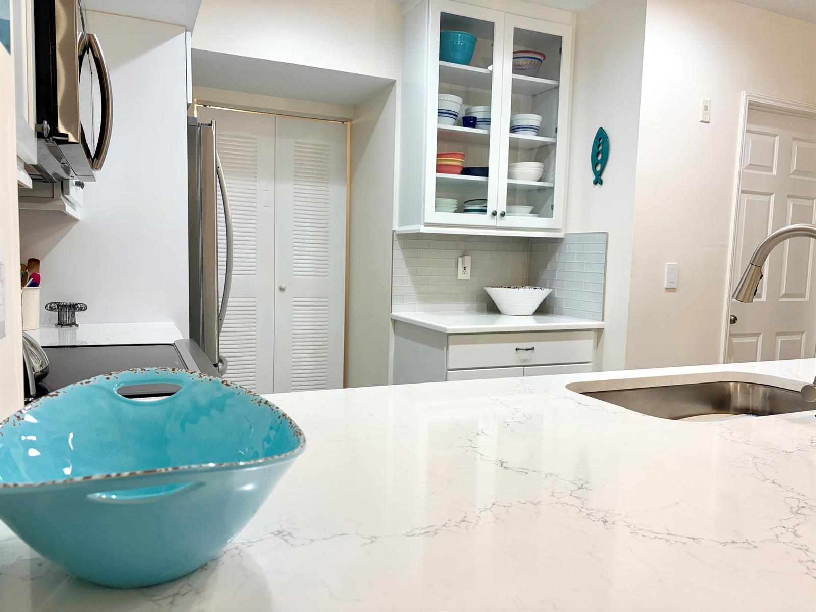 A bright kitchen features white cabinets, quartz countertops, a stainless steel sink, and glass-front shelving — 242 Village Boulevard # 2304, Tequesta FL 33469, TEQUESTA, FL