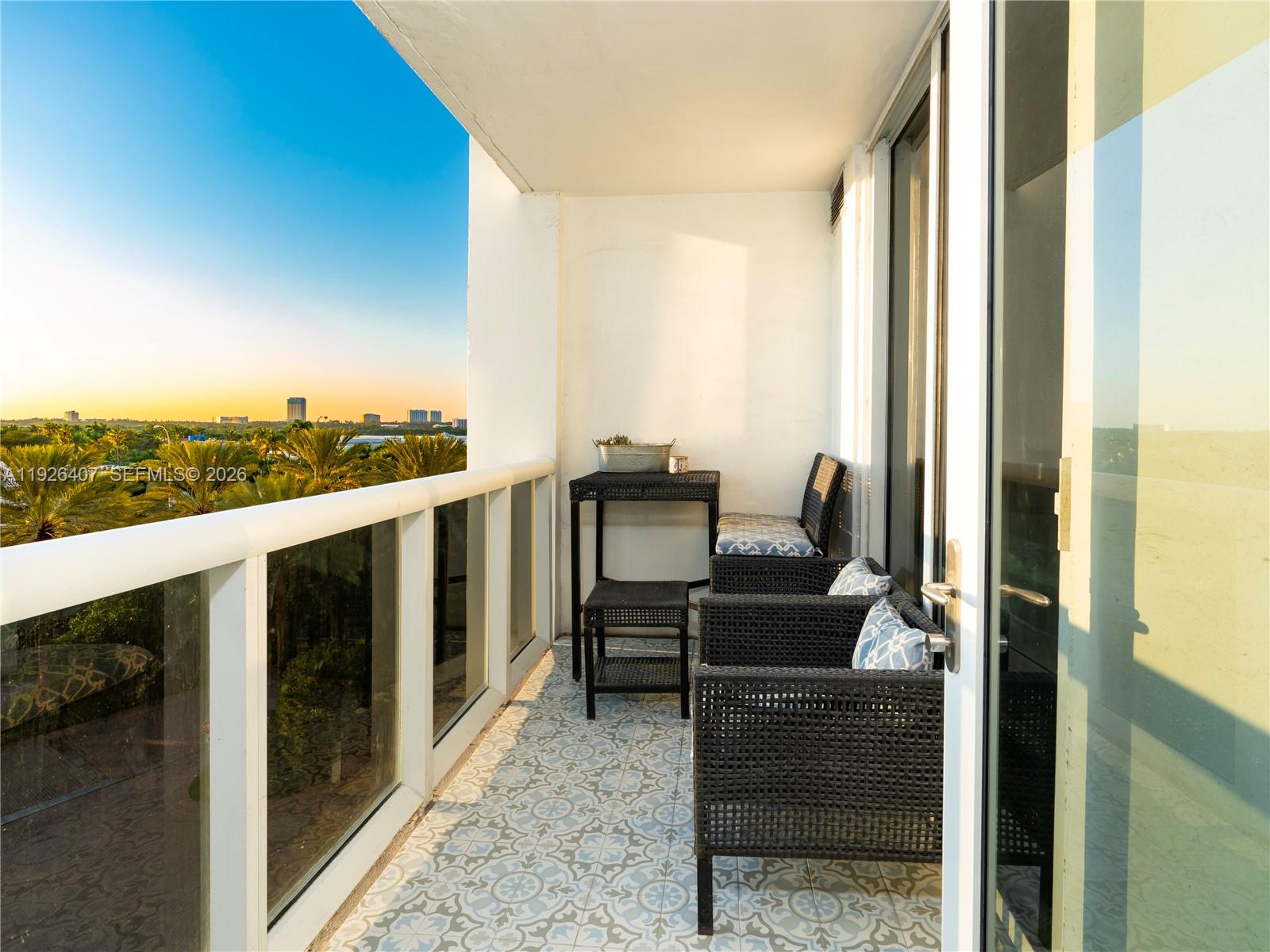 A balcony features patterned tile flooring, wicker furniture, and a glass railing overlooking palm trees — 10275 Collins Ave # 527, Bal Harbour FL 33154, BAL HARBOUR, FL
