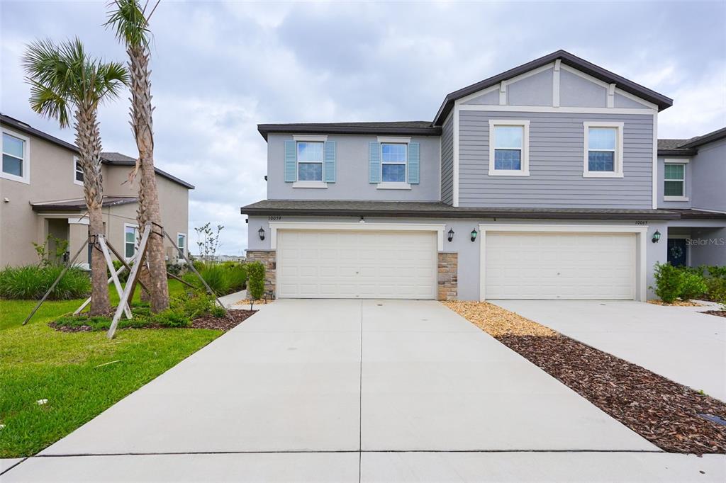 A two-story duplex features light gray siding, two white garage doors, and a concrete driveway — 10059 TRUMPET HONEYSUCKLE WAY, SAN ANTONIO, FL