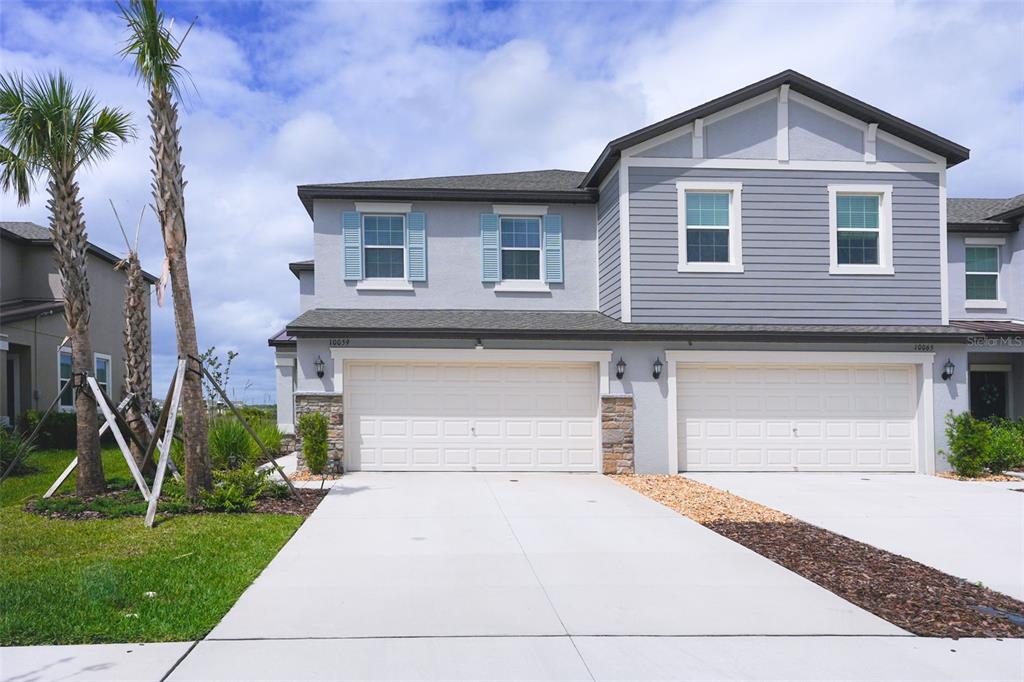 A two-story duplex with gray siding, stone accents, and two side-by-side garage doors — 10059 TRUMPET HONEYSUCKLE WAY, SAN ANTONIO, FL