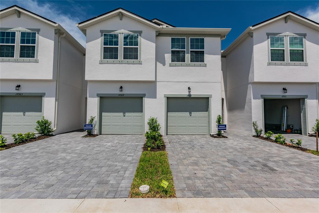 A row of four attached townhouses with light-colored stucco facades, green garage doors, and paved driveways — 19309 BLUE POND DRIVE, LUTZ, FL