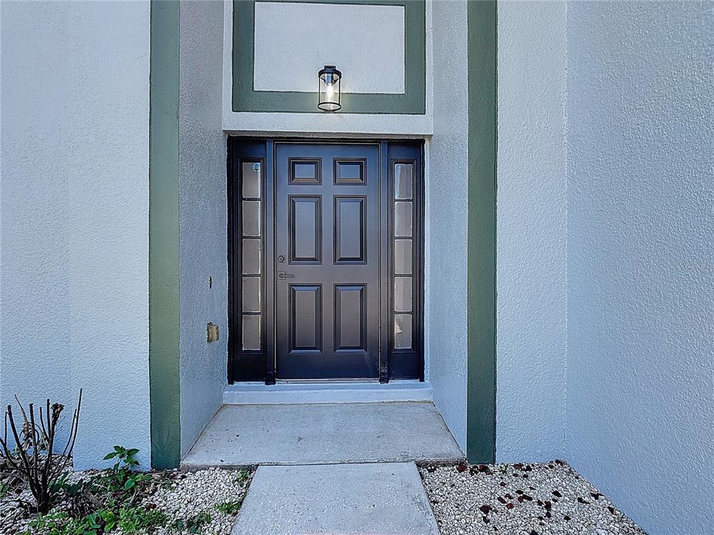A dark front door with side windows is centered on a light blue exterior wall — 965 ALSACE DRIVE, KISSIMMEE, FL