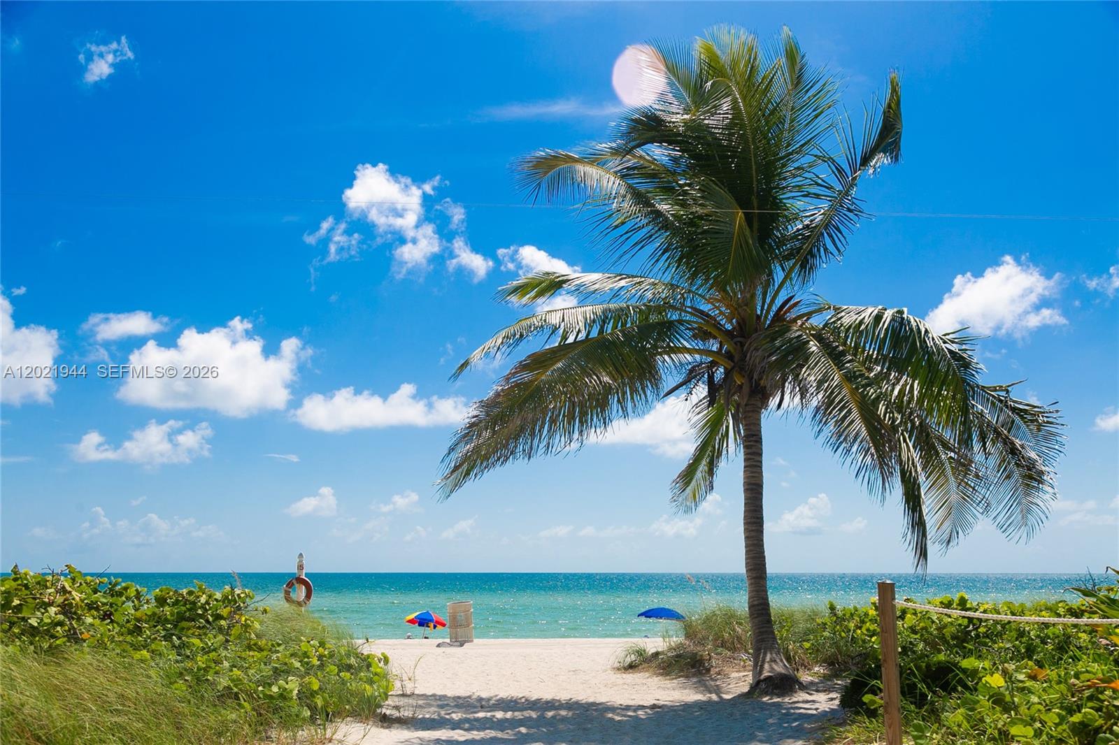 A sandy beach features two umbrellas overlooking the blue ocean under a clear sky — 8855 Collins Ave # 6F, Surfside FL 33154, SURFSIDE, FL